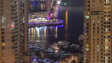 Dubai Marina 'daki gezinti güvertesi ve kanalı. Gece vakti, Birleşik Arap Emirlikleri civarında aydınlık lüks gökdelenleri var. Yatlar ve tekneler yukarıdan kanala bakıyor.