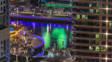 Dubai Marina 'daki gezinti güvertesi ve kanalı. Gece vakti, Birleşik Arap Emirlikleri civarında aydınlık lüks gökdelenleri var. Yatlar ve restoranlarda oturan insanlar yukarıdan hava manzaralı.