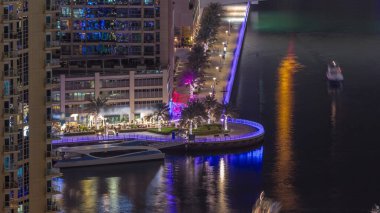 Yürüyen insanlarla ve Dubai Marina 'da kanal gezintisi. Gece vakti aydınlatılmış restoranlar, Birleşik Arap Emirlikleri. Yatlar ve tekneler yukarıdan kanala bakıyor.