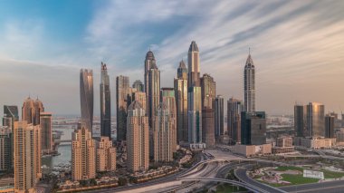 Dubai Marina 'nın gökdelenleri. Sabah saatlerinde aydınlatılmış en yüksek konut binaları. Otoyol kavşağında trafik var. Güneş doğarken JLT bölgesinin hava üst görüntüsü