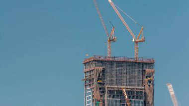 Yüksek çok katlı bina yapım aşamasında ve birçok vinç zaman ayarlı. Arka planda mavi gökyüzü olan yeni gökdelenin şantiyesinde aktif iş.
