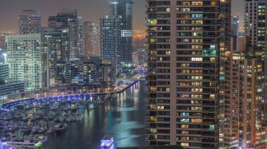 Dubai marinasına giden hava manzaralı, gece saatlerinde yüzen yatlarla kanalın etrafında parlayan pencereleri olan gökdelenler. Yat kulübünde beyaz tekneler var.