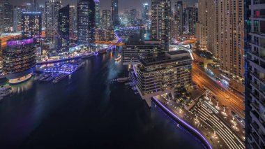 Dubai marinasına giden hava manzaralı gökdelenler ve kanalın etrafındaki JBR kuleleri gece saatlerinde yüzen yatlarla aydınlatıldı. Yat kulübünde beyaz tekneler var.