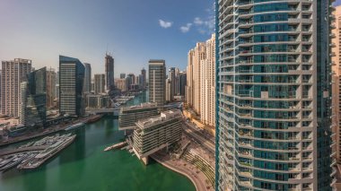 Dubai marinasına ve kanalın etrafındaki JBR gökdelenlerine yüzen tekneler panoramik zaman çizelgesiyle hava görüntüsü. Pencereleri ve balkonları olan uzun cam kuleler. Yat kulübünde beyaz tekneler var.