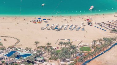 Güneşin ve denizin tadını çıkaran turistlerle dolu kalabalık JBR plajı, yukarıdan şemsiyeli hava manzarası altında oturuyordu. Sarı kum ve palmiyeler etrafta, kayıklar suda yüzüyor.