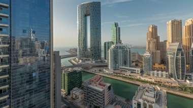 Panorama, Dubai Marina gökdelenlerini ve JBR bölgesini lüks binalar ve tatil köylerinin zaman çizelgeleriyle gösteriyor. Palmiyeler ve kanalda yüzen teknelerle dolu bir liman.