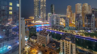 Dubai Marina gökdelenleri ve JBR bölgesi güneş doğmadan önce aydınlanmış lüks binalar ve gece gündüz geçiş zamanları ile. Palmiyeler ve kanalda yüzen teknelerle dolu bir liman.