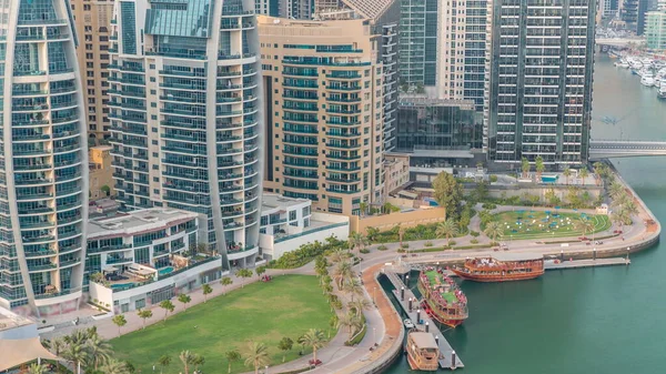 Rıhtım kenarındaki eski ahşap gemi, palmiyeler ve yeşil çimenli havadan zaman ayarlı. Dubai Marina 'daki Dhow gezisinde gökdelenler, Birleşik Arap Emirlikleri