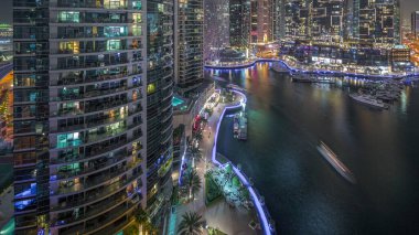 Dubai Marina hava sahasında palmiyelerle rıhtım gezintisi. Yatlar ve tekneler suyun üzerinde yüzüyor. Parlak pencereli gökdelenler
