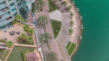 Dubai Marina 'da avuçiçi avuçlarla rıhtım gezintisi. Ağaçların yanındaki restoran masaları ve sandalyeleri. Yatlar ve tekneler suda yüzüyor.