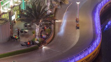 Dubai Marina hava sahasında palmiyelerle rıhtım gezintisi. İnsanlar bir bankta yürüyor ve oturuyor. Yatlar ve tekneler suda yüzüyor.