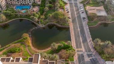 Köprüsü olan gölet ve Yeşiller Bölgesi hava zaman diliminde alçak binalar. Palmiyeler ve ağaçlarla dolu Dubai silueti. Gölgeler hızlı hareket ediyor