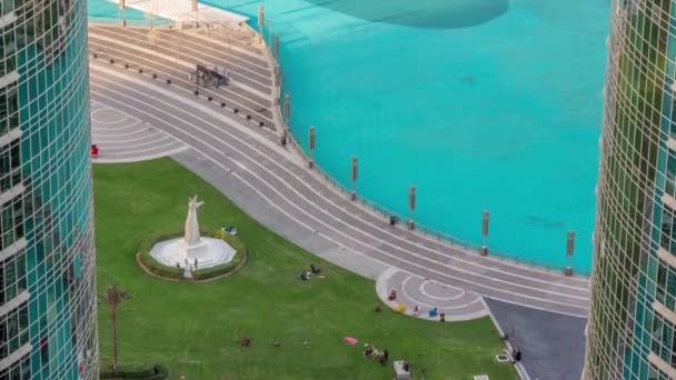Park se zeleným trávníkem u fontán vzdušného času. Věže kolem umělého jezera v centru Dubaje. — Stock video