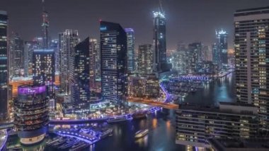 Kanal çevresindeki Dubai marina gökdelenlerine gece vakti yüzen teknelerle hava görüntüsü.