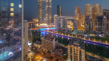 Dubai Marina gökdelenleri ve JBR bölgesi güneş doğmadan önce aydınlanmış lüks binalar ve gece gündüz geçiş zamanları ile. Palmiyeler ve kanalda yüzen teknelerle dolu bir liman.