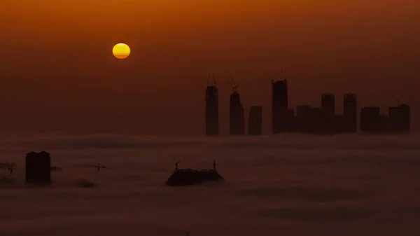 高層ビルや塔が朝の霧の空中のタイムラプスで覆われているドバイクリークハーバーの日の出 ビルやオレンジの空の後ろに大きな赤い太陽が昇る — ストック写真