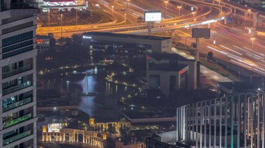 Dubai Internet City Gölü 'nün havadan görünüşü. Çeşme ve binalar gece vakti yukarıdan görünüyor. Etrafta palmiye ağaçları ve otoyolda trafik