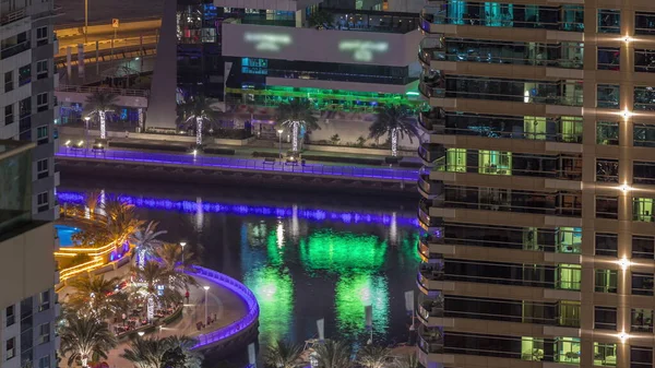 Dubai Marina 'daki gezinti güvertesi ve kanalı. Gece vakti, Birleşik Arap Emirlikleri civarında aydınlık lüks gökdelenleri var. Yatlar ve restoranlarda oturan insanlar yukarıdan hava manzaralı.