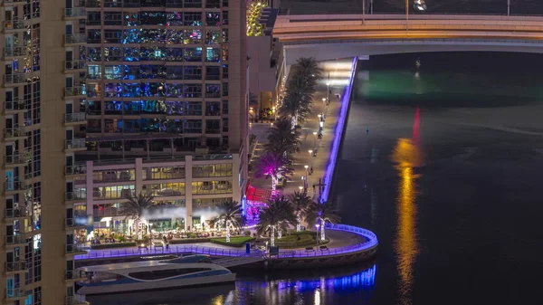 Yürüyen insanlarla ve Dubai Marina 'da kanal gezintisi. Gece vakti aydınlatılmış restoranlar, Birleşik Arap Emirlikleri. Yatlar ve tekneler yukarıdan kanala bakıyor.