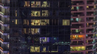 Modern ofisteki pencere ışıkları ve gece vakti balkonlu binalar. İçinde aydınlık odalar olan çok katlı gökdelenler.