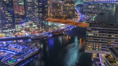 Dubai marinasının rıhtım manzarası ve kanalın etrafındaki gökdelenleri gece saatlerinde yüzen yatlarla aydınlatıyor. Yat kulübünde beyaz tekneler var.