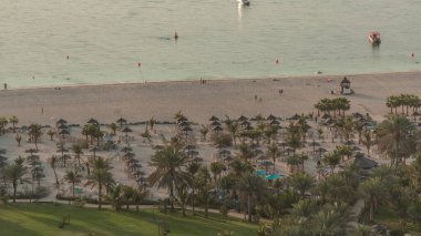 JBR 'deki kumsalda, deniz kenarındaki havadan kumsala yakın bir yerde. Palmiyeli yeşil çimenler ve şemsiyeli sandalyelerde dinlenen insanlar.