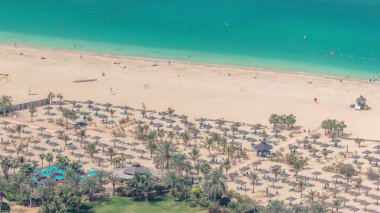 Güneşin ve denizin tadını çıkaran turistlerle dolu kalabalık JBR plajı, yukarıdan şemsiyeli hava manzarası altında oturuyordu. Etrafta yeşil çimenler ve palmiyeler, suyun üzerinde yüzen kayıklar.