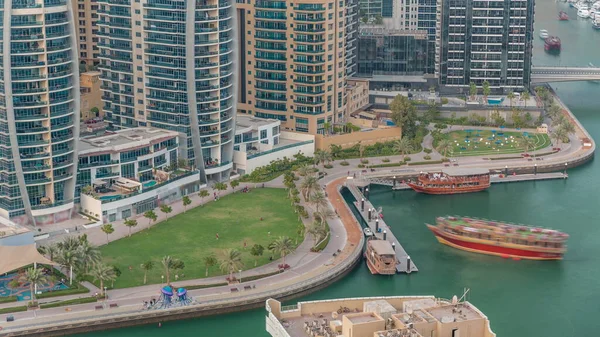 Rıhtım kenarındaki eski ahşap gemi, palmiyeler ve yeşil çimenli havadan zaman ayarlı. Dubai Marina 'daki Dhow gezisinde gökdelenler, Birleşik Arap Emirlikleri
