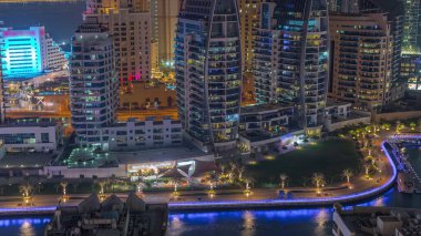 Dubai Marina gökdelenleri ve lüks binaları olan JBR bölgesi ve gece uçuşları için zaman çizelgesi. Palmiyeler, yeşil çimenler ve kanalda yüzen teknelerle aydınlatılmış liman.