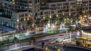 Dubai şehir merkezindeki yolda yoğun bir trafik var. Hava gece saatleri. Palmiyeler ve restoranlarla dolu bir bulvar. Birçok araba karşıya geçiyor, BAE