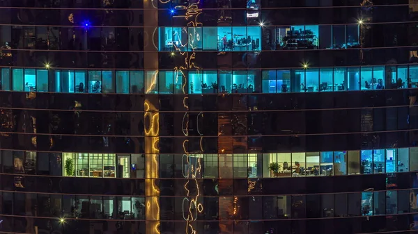 Office windows of a glazed skyscraper glow at night with city lights reflection aerial timelapse. Illuminated tower at evening