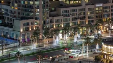 Dubai şehir merkezindeki yolda yoğun bir trafik var. Hava gece saatleri. Palmiyeler ve restoranlarla dolu bir bulvar. Birçok araba karşıya geçiyor, BAE