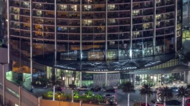 Yüksek sınıf bir binanın dairelerinde gece vakti aydınlatılmış panoramik pencereler. Girişte park yeri olan gökdelende parlayan ışıklar.