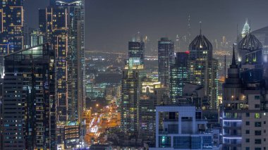 Dubai Marina ve Medya Şehri bölgelerinde modern gökdelenler ve ofis binaları gece uçuşları. Üst geçitte trafik ve şehir merkezindeki kulelerde kavşak.
