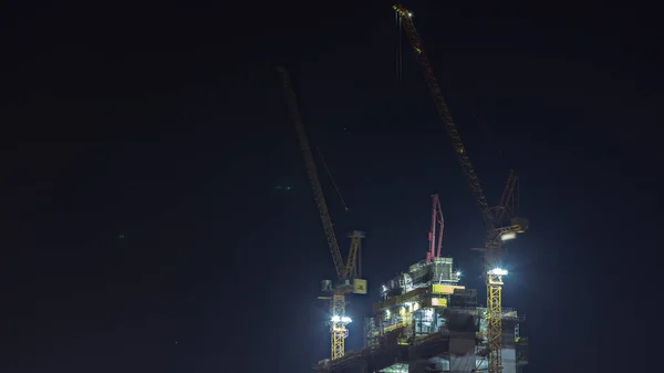 Skyscraper under construction with cranes night timelapse. Building of new multi-storey tall tower with builders working on top