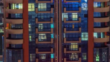 Aydınlatılmış çalışma odası saatleriyle apartman penceresi ön cephesinin gece görüşü. Gökdelende parlayan ışık ve cam yüzey