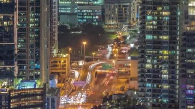 Dubai Marina ve Medya Şehri bölgelerinde modern gökdelenler ve ofis binaları gece uçuşları. Yeşil park yakınlarındaki üst geçitte trafik ve kavşak.