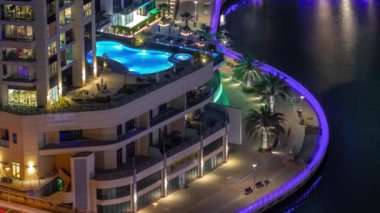 Dubai Marina rıhtımı ve şehir güvertesi gece vardiyası yukarıdan. Kanallar boyunca gökdelenlerin havadan görüntüsü. Çatıda aydınlatılmış bir yüzme havuzu.