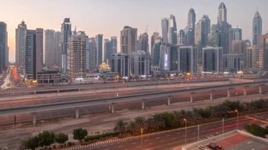 Dubai marinası gökdelenlerin en uzun bloğu günden geceye geçiş zamanı. JLT bölgesinden apartman binalarına, otellere ve otoyol ve metro hattı yakınlarındaki iş kulelerine hava panoramik görüntüsü.