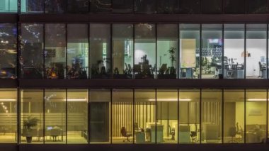 Modern ofis binalarındaki büyük parlayan pencereler geceleri zaman ayarlı, sıra sıra parlıyor. İnsanlar içeride masalarda çalışıyor.
