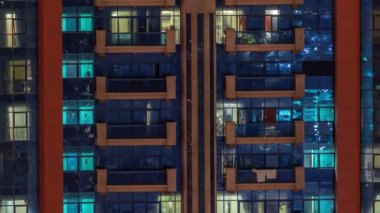 Aydınlatılmış çalışma odası saatleriyle apartman penceresi ön cephesinin gece görüşü. Balkonlu gökdelende parlayan ışık