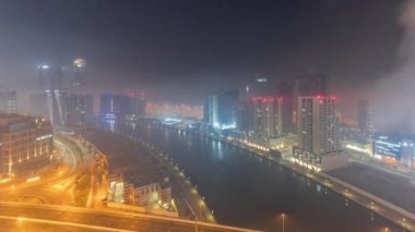 Tüm gece boyunca Business Bay bölgesinde modern şehir mimarisi. Dubais 'in panoramik hava manzarası gökdelenleri aydınlatırken zaman kavramları yol trafiğiyle çakışıyor. Sisli hava ve gün doğumu