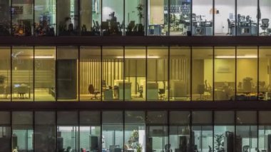 Modern ofis binalarındaki büyük parlayan pencereler geceleri zaman ayarlı, sıra sıra parlıyor. İnsanlar içeride masalarda çalışıyor.