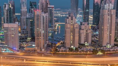 Dubai Marina 'nın gökdelenleri gece gündüz geçiş zamanlarında en yüksek konut binalarıyla aydınlandı. Güneş doğmadan önce JLT bölgesinin havadan görüntüsü. Yatlar suda yüzüyor.
