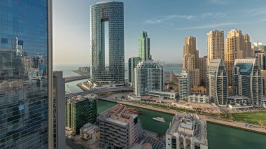 Panorama, Dubai Marina gökdelenlerini ve JBR bölgesini lüks binalar ve tatil köylerinin zaman çizelgeleriyle gösteriyor. Palmiyeler ve kanalda yüzen teknelerle dolu bir liman.