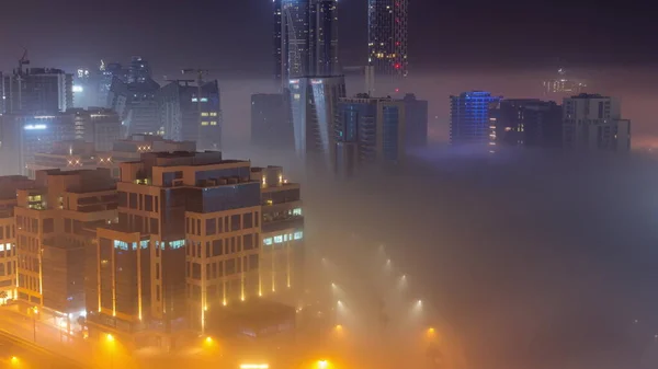 Buildings are covered in thick layer of fog in Business Bay night timelapse. Illuminated skyscrapers around water canal aerial top view