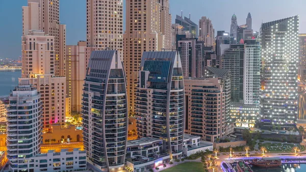 Dubai Marina skyscrapers and JBR district before sunrise with illuminated luxury buildings and resorts aerial night to day transition timelapse. Waterfront with palms and green lawn
