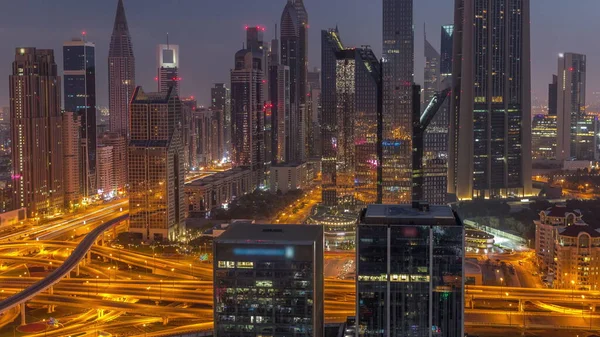 Panorama of Dubai Financial Center district with tall skyscrapers with illumination night to day transition timelapse. Aerial view to towers along busy highway before sunrise