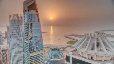 JBR ve Dubai Marina gökdelenlerinin hava günbatımı manzarası ve yukarıdan gelen lüks binalar. Turuncu gökyüzünde kumsalı ve güneşi olan modern kuleler denize yansıyor.