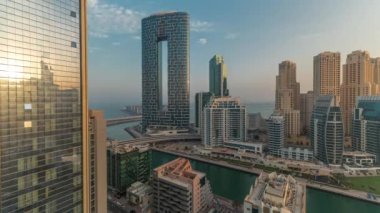 Güneş doğarken Dubai Marina gökdelenleri ve JBR semti lüks binalardan ve tatil beldelerinden yansıyan güneşle birlikte. Palmiyeler ve kanalda yüzen teknelerle dolu bir liman.